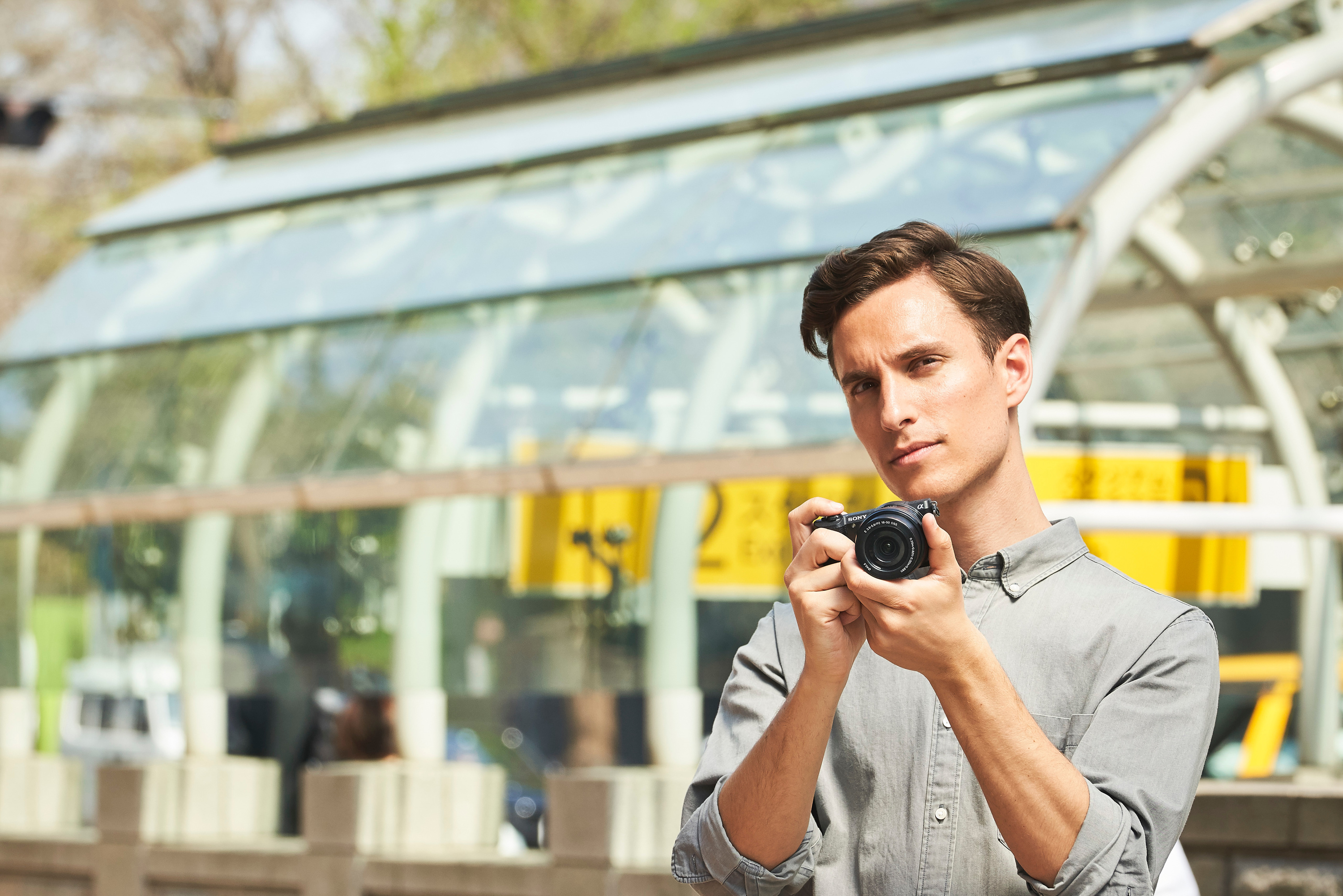Sony A5100 -kompaktikamera Mies seisoo kaarevan lasikatoksen edessä ja pitelee kädessään Sony A5100 -kompaktikamera.a