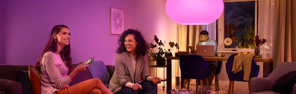 Philips Hue - Silta vai Bluetooth? Kaksi naista istuu sohvalla violetin värisen Philips Hue -lampun vieressä. Taka-alalla mies työskentelee tietokoneella pöydän ääressä.