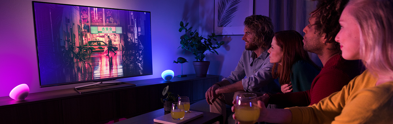Viihdekäyttöön Kaksi pariskuntaa istumassa sohvalla katselemassa TV:tä, jonka vieressä kaksi Philips Hue -valoa.