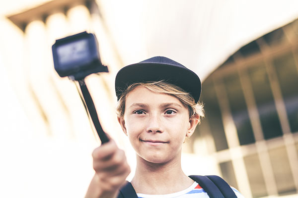 Käytössä monipuolinen Poika, jolla musta lippalakki päässään kuvaa itseään selfiekeppiin asennetulla action-kameralla