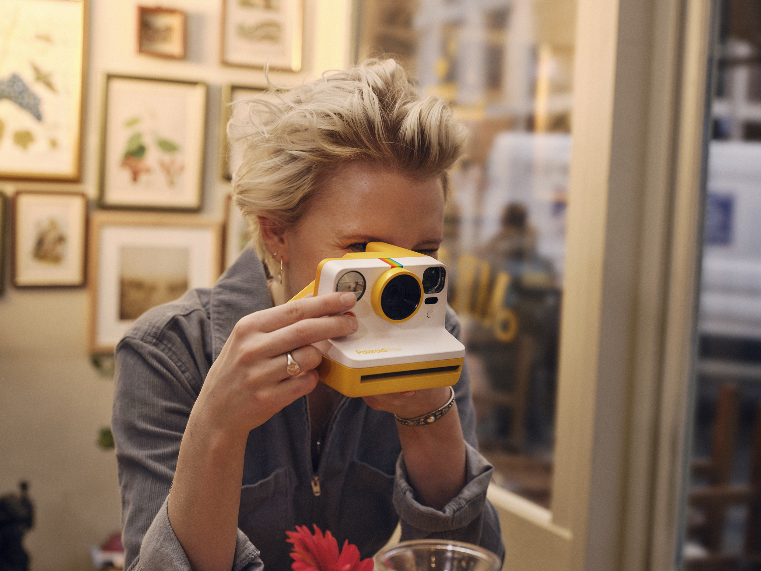 Polaroid NOW Nainen pitelee oranssia Polaroid NOW -kameraa kasvojensa edessä