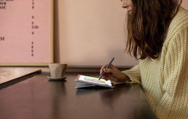 Surface Go 2:ssa on 10,5 tuuman kosketusnäyttö, mikä tekee surffailusta miellyttävää Nainen istuu kahvilla, hänellä on Surface Pen -kynä kädessään ja Surface Go 2 edessään
