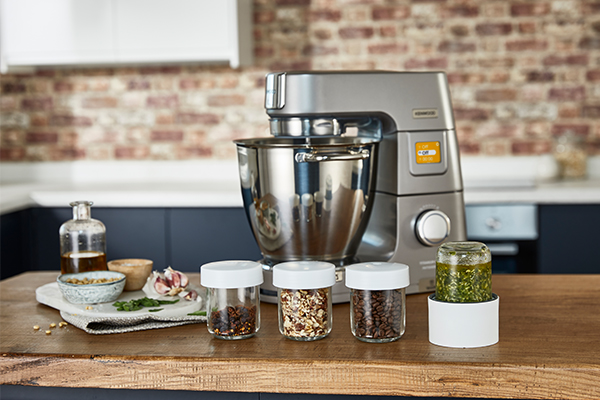 Kenwood Teräksen värinen Kenwood-yleiskone keittiön työtasolla, jonka edessä yrttejä muovipurkeissa