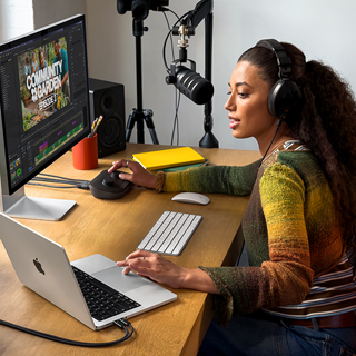 Nainen työskentelee kotonaan työpöydän ääressä MacBook Prolla, joka on liitetty ulkoiseen näyttöön; naisella on kuulokkeet, ja hän puhuu mikrofoniin