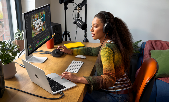 Nainen työskentelee kotonaan työpöydän ääressä MacBook Prolla, joka on liitetty ulkoiseen näyttöön; naisella on kuulokkeet, ja hän puhuu mikrofoniin