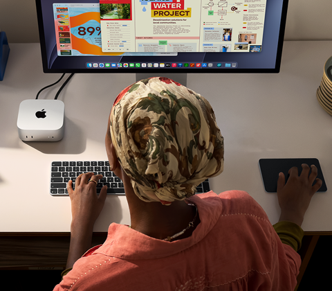 Henkilö kirjoittaa työpöydän ääressä Mac minillä, Studio Display liitettynä, Magic Keyboard, Magic Trackpad, erilaisia esineitä