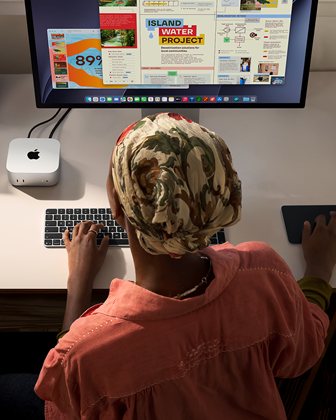 Henkilö kirjoittaa työpöydän ääressä Mac minillä, Studio Display liitettynä, Magic Keyboard, Magic Trackpad, erilaisia esineitä