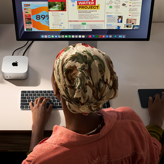 Henkilö kirjoittaa työpöydän ääressä Mac minillä, Studio Display liitettynä, Magic Keyboard, Magic Trackpad, erilaisia esineitä