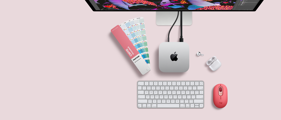 Mac mini ylhäältä, hopeanväristä alumiinia, pyöristetyt kulmat, kiiltävä musta Apple-logo keskellä, liitetty johdolla, Studio Display osittain esillä sen päällä, AirPods 4 ‑kotelo avoinna, Pantone-värinäytteitä sekä punainen Logitech-näppäimistö ja -hiiri