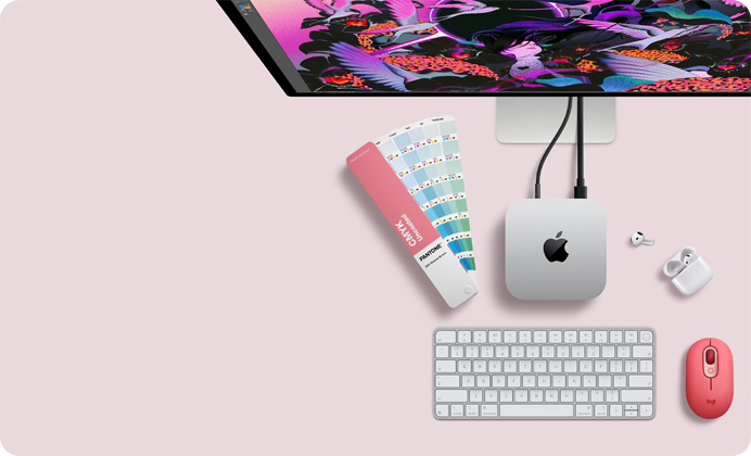 Mac mini ylhäältä, hopeanväristä alumiinia, pyöristetyt kulmat, kiiltävä musta Apple-logo keskellä, liitetty johdolla, Studio Display osittain esillä sen päällä, AirPods 4 ‑kotelo avoinna, Pantone-värinäytteitä sekä punainen Logitech-näppäimistö ja -hiiri