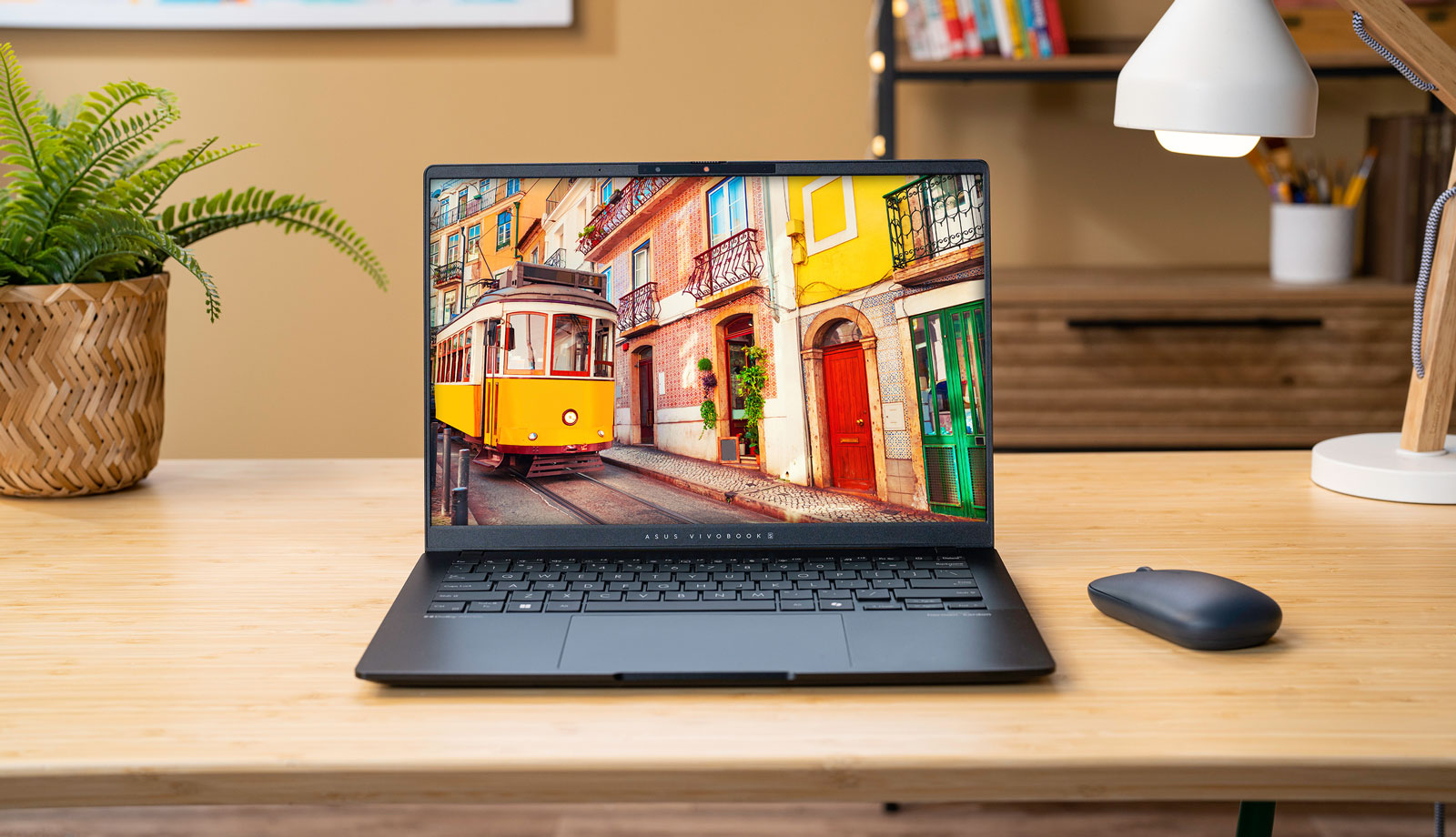 Pöydällä oleva Asus Vivobook S 14 M5406 OLED kannettava tietokone ja hiiri, lamppu ja viherkasvi sen vieressä