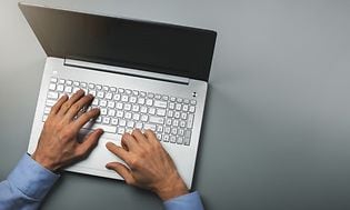 Kädet kirjoittamassa tietokoneella harmaalla pöydällä
