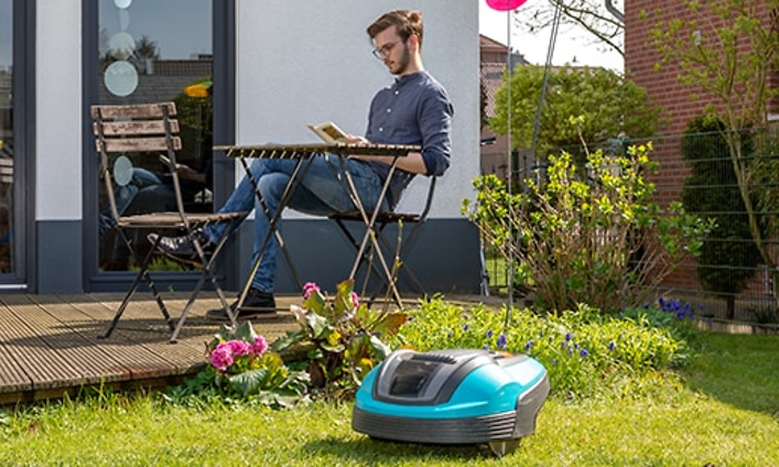 Mies istuu lukemassa puutarhassa sillä aikaa kun robottiruohonleikkuri leikkaa nurmikkoa