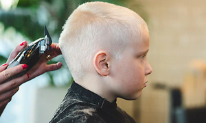 Poika kuvattuna sivusta päin. Takana seisoo nainen hiustenleikkuukoneen kanssa