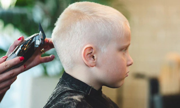 Poika kuvattuna sivusta päin. Takana seisoo nainen hiustenleikkuukoneen kanssa