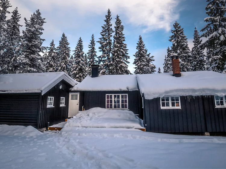 Tumma mökki lunta katolla ja maassa, taustalla sininen taivas ja kuusia