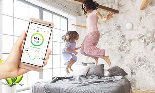 Airthings WavePlus ja puhelin Airthings-sovelluksella makuuhuoneessa, jossa äiti ja tytär hyppäävät sängyllä