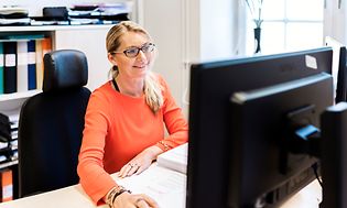 Hymyilevä nainen istuu työskentelemässä tietokoneella työpöydän ääressä
