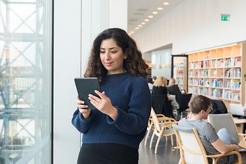 Nainen seisoo kirjastossa ikkunan ääressä lukemassa jotain tabletiltaan