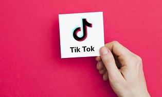 Käsi pitelee valkoista korttia, jossa on TikTok-logo ja takana vaaleanpunainen tausta
