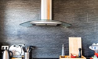 Teräksen värinen seinäkiinnitteinen liesituuletin keittiön harmaata seinää vasten keittotason yläpuolella