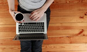 Mies istuu lattialla kannettava sylissään ja pitelee kahvikuppia toisessa kädessään