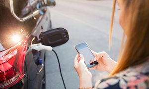 Nainen pitelee älypuhelinta sähköauton vieressä
