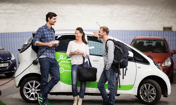 Kaksi miestä ja nainen seisovat valkoisen sähköauton vieressä