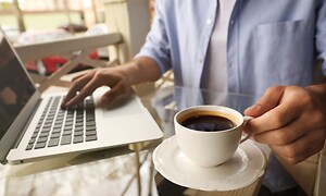 Tietokoneet - Korjaus - Henkilö juo kahvia kannettavan tietkoneen vieressä