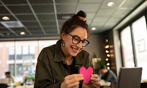 Hymyilevä nainen istuu kannettavan äärellä ja pitelee pinkkiä sydäntä kädessään