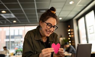 Hymyilevä nainen istuu kannettavan äärellä ja pitelee pinkkiä sydäntä kädessään