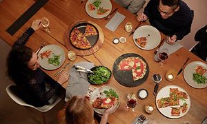 Joukko ruokailijoita nauttii pizzaa suuren puupöydän ääressä