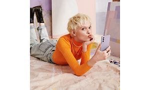 Nainen makaa lattialla ja pitelee liilan väristä Samsung Galaxy S21 FE -puhelinta kädessään