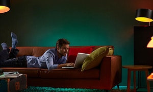 Mies makaa sohvalla ja käyttää kannettavaa tietokonetta eri lamppujen hallitsemiseen ympärillään