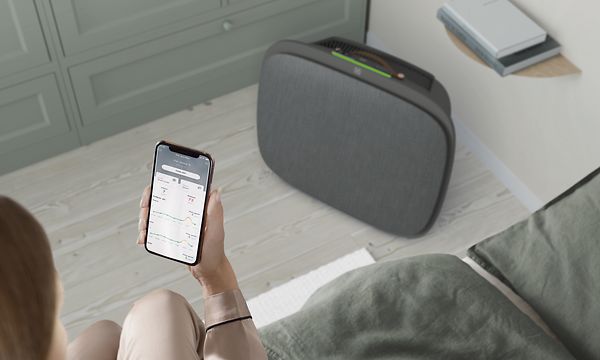 Tummanharmaa Well 5 -ilmanpuhdistin lattialla ja Wellbeing-sovellus naisen puhelimessa