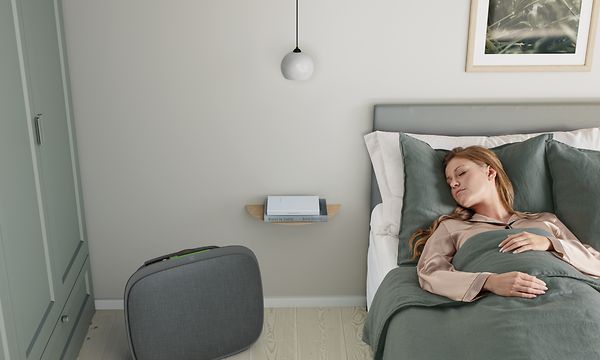 Nainen nukkuu sängyssä ja tummanharmaa Well 5 -ilmanpuhdistin lattialla sängyn vieressä