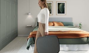 Makuuhuoneessa oleva nainen kantaa tummanharmaata Well 7 -ilmanpuhdistinta kuin matkalaukkua