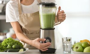 Tehosekoittimessa vihreää smoothieta ja nainen säätämässä nopeutta