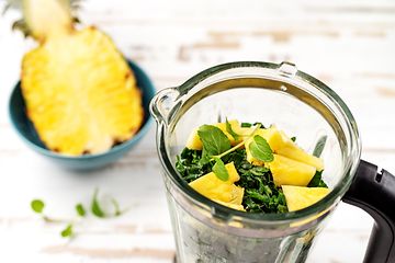 Tehosekoitin, jossa on yrttejä ja leikattuja ananaspaloja sekä puolikas ananas tehosekoittimen vieressä
