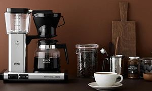 Moccamaster-kahvinkeitin, kahvikuppi ja lasipurkillinen kahvipapuja pöydällä