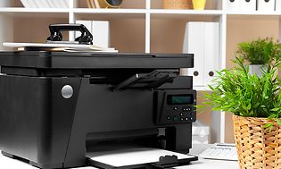 Musta tulostin toimiston pöydällä, jonka vieressä viherkasvi ja takana kansioita hyllyssä