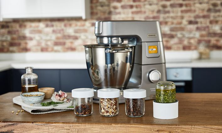 Teräksen värinen Kenwood-yleiskone keittiön työtasolla, jonka edessä erilaisia mausteita purkeissa