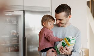 Jääkaapin edessä seisova isä ja tyttölapsi sylissä sekä maitopurkki