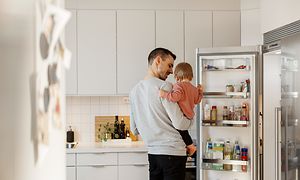 Avoimen jääkaapin edessä seisova isä ja tyttölapsi sylissään