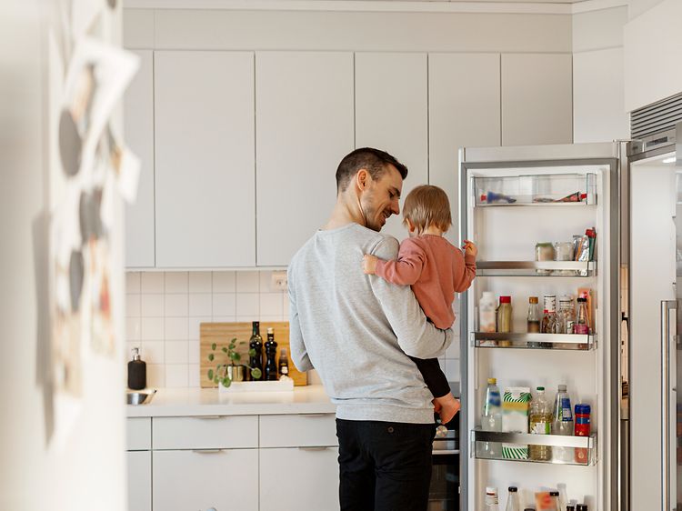 Avoimen jääkaapin edessä seisova isä ja tyttölapsi sylissään