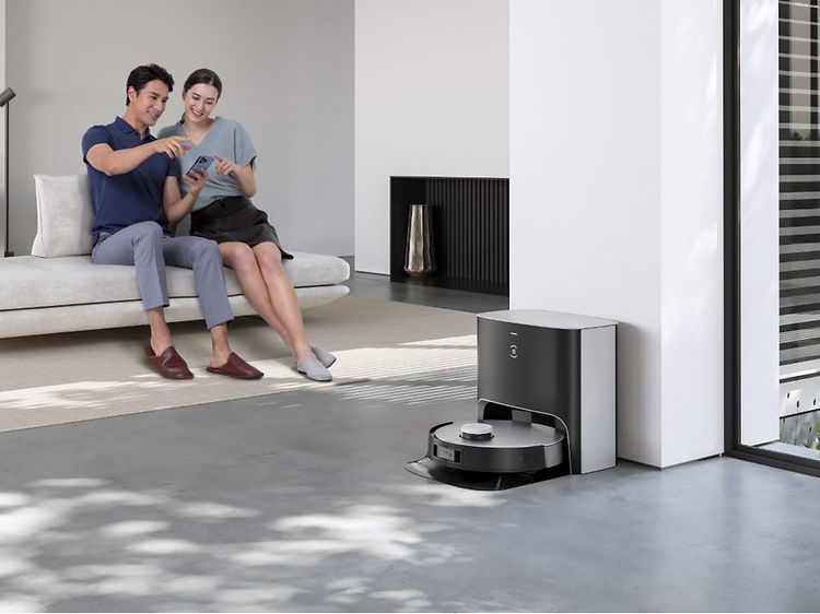 Latauksessa oleva Ecovacs Deebot X1 Plus -robotti-imuri ja taustalla sohvalla istuva pariskunta