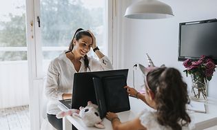 Nainen työskentelee kannettavalla pöydän ääressä tyttärensä vieressä, joka käyttää tablettia