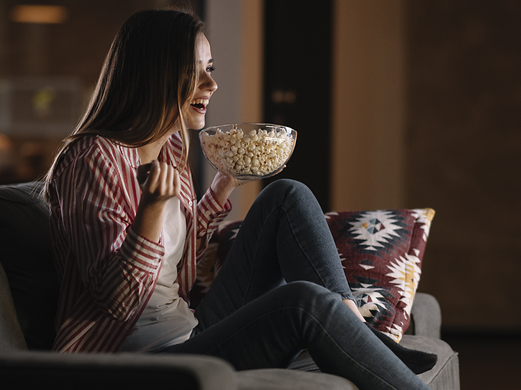 Nuori nainen istuu sohvalla katsomassa televisiota popcorn-kulho kädessään