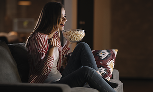 Nuori nainen istuu sohvalla katsomassa televisiota popcorn-kulho kädessään