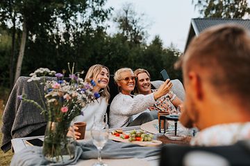 Kaksi naista ja mies istuvat vierekkäin ulkona puutarhassa ja ottavat selfie-kuvaa itsestään